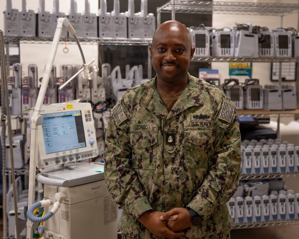 Military man smiles next to medical ventilator and equipment.